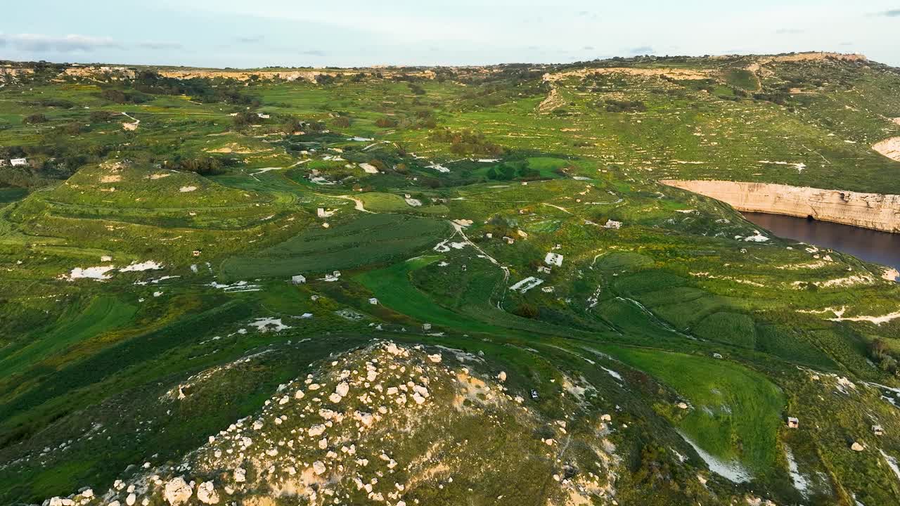 Aerial drone footage captures vibrant green terraced fields and dramatic limestone cliffs of Miġra l-Ferħa, Malta. The view highlights the natural beauty of this stunning Mediterranean landscape.