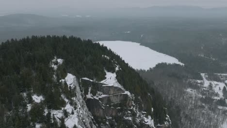 Hombre-En-La-Cima-De-La-Montaña-Rocosa-En-La-Nieve-Con-Bosque-De-Pinos-Durante-El-Invierno-En-Quebec,-Canadá