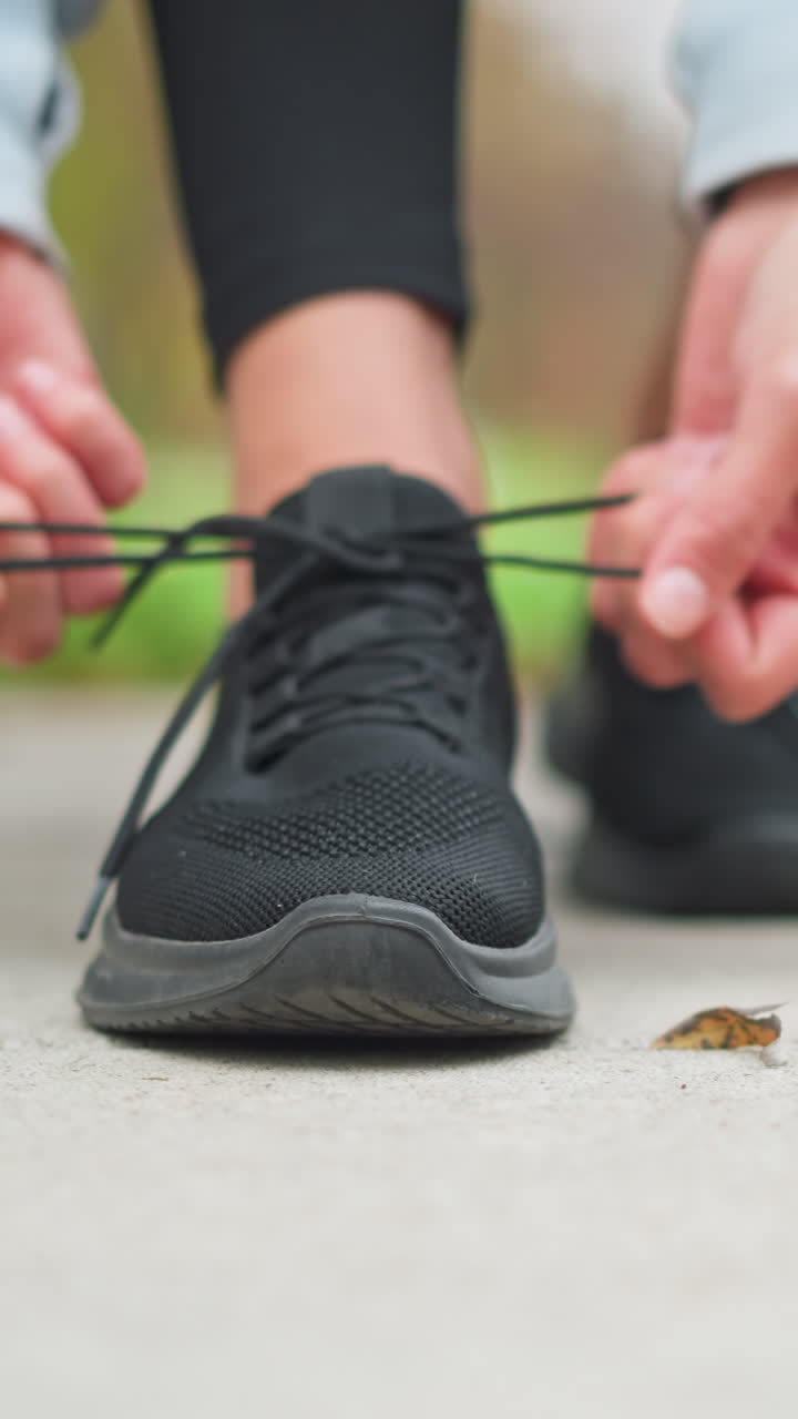 primo piano di una donna che allaccia i lacci delle sue scarpe da ginnastica nere con l'attenzione sulle mani, che mostrano anelli, preparandosi per l'esercizio, allenamento all'aperto in un parco in una giornata di sole, indossando abbigliamento sportivo