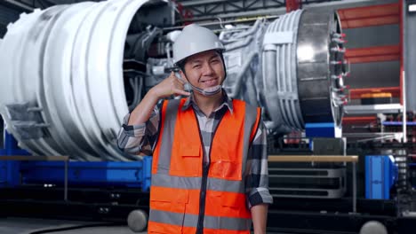 asian male engineer with safety helmet smiling to camera and making call me gesture while standing with airplane engine maintenance conducted, analytics checking the turbine