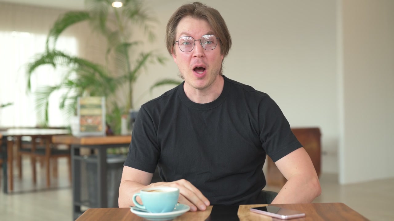 Portrait of Scandinavian Northern European man sitting in coffee shop cafe restaurant vlogging and talking to camera happily