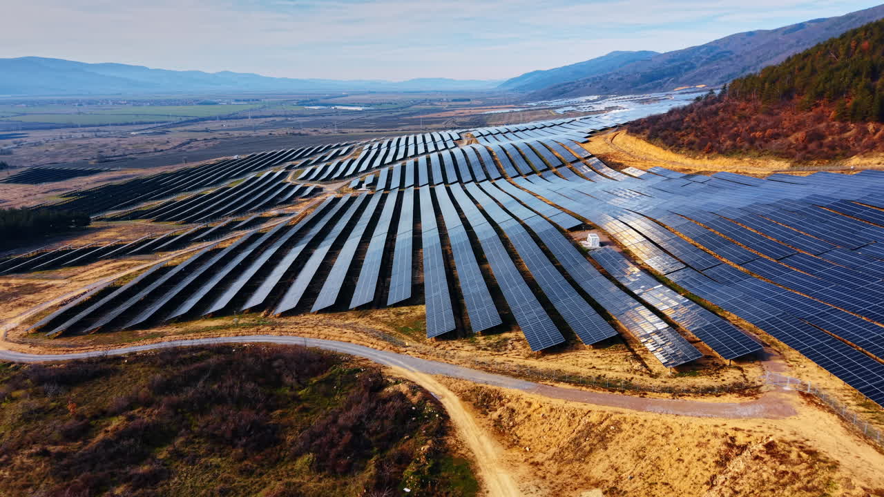 Luftfoto af solcellepark i bjergdal. Fotovoltaiske moduler arrangeret i rækker på bakket terræn under blå himmel