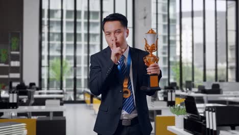 asian business man in a suit and tie with a gold medal and trophy making shh gesture telling it is a secret to win as the first winner in office