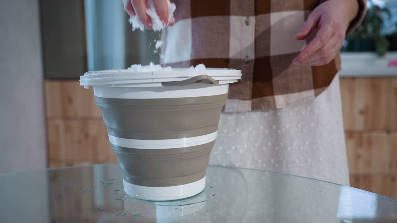 Serene gardener lifts icy bucket full of melting snow onto glass table in cozy indoor wood paneled room capturing gentle drips and light reflections in slow mindful ritual under soft light