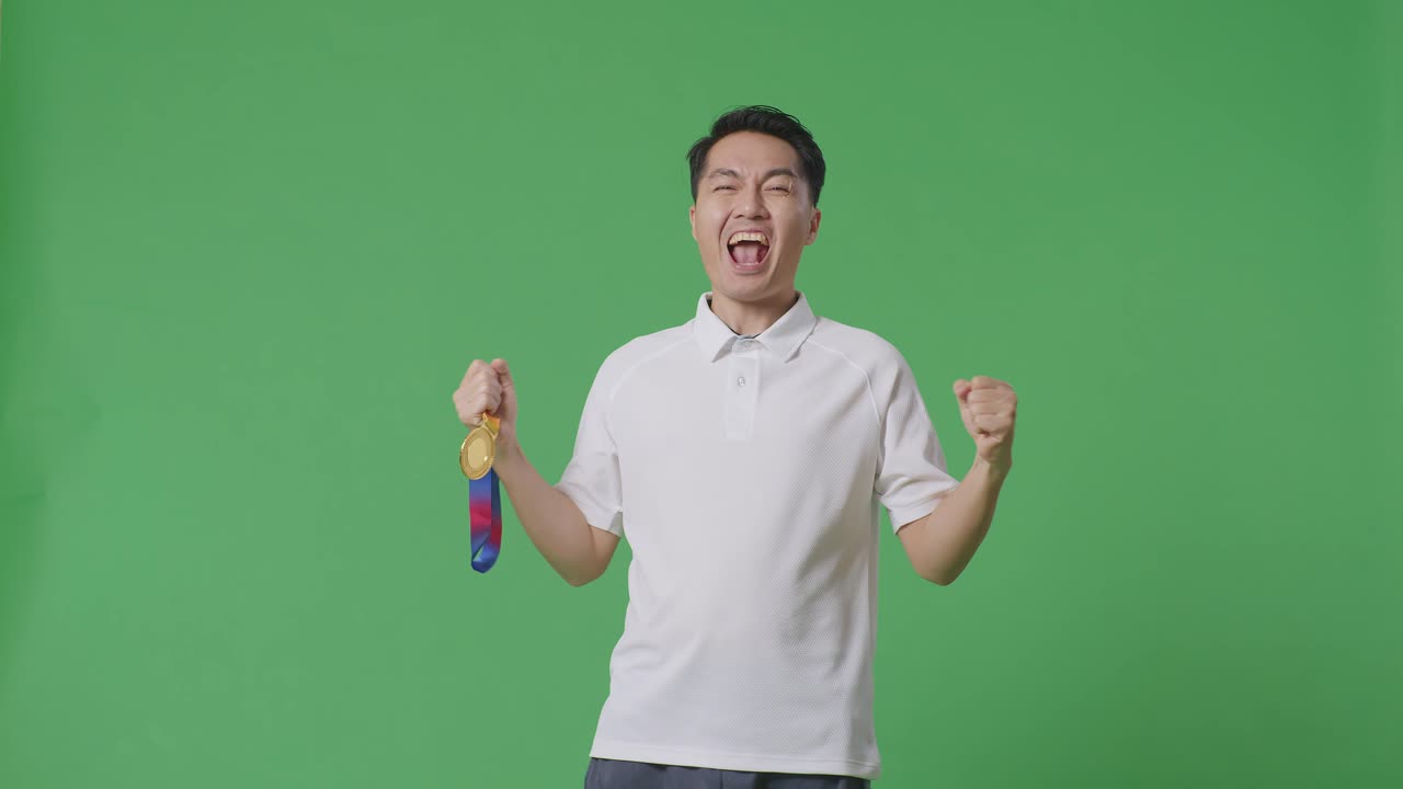 금메달을 바라보는 아시아인 남자가 골을 외치며 우승을 축하하며 스튜디오의 녹색 스크린 배경에서 첫 번째 우승자로 승리합니다.