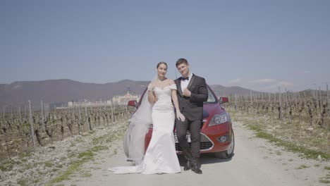 pareja de bodas posando por un coche rojo en un viñedo