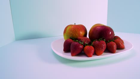 Fresh-big-red-tasty-ripe-strawberries-and-red-apples-covered-by-water-droplets-rotates-slowly-on-a-white-plate-on-light-blue-background,-healthy-food-concept,-medium-shot,-camera-rotate-left