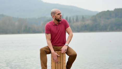Pensive-musician-playing-cajon-in-nature-against-lake-and-mountains
