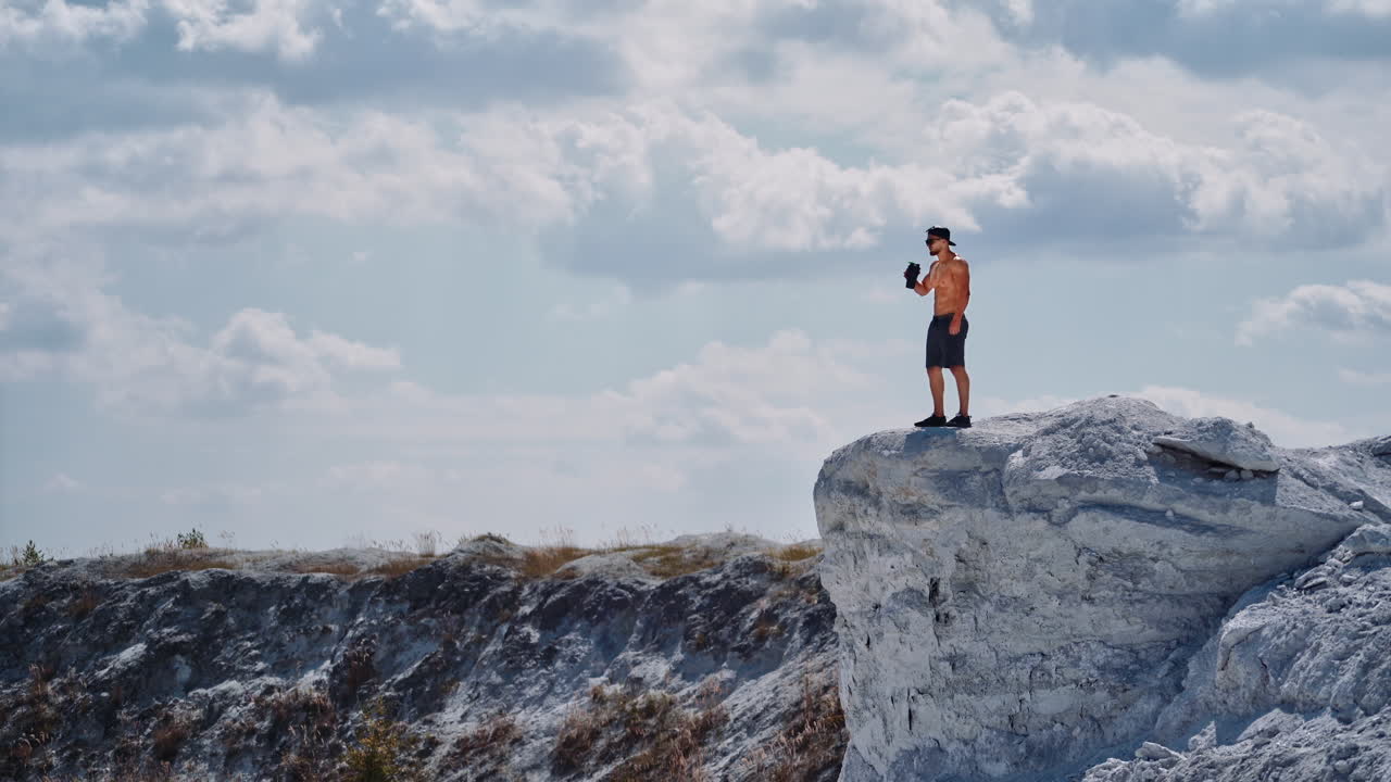 Athlete drinking water on a white rock in summer. Shirtless man with flask in white canyon in a sunny day. Healthy sportsman after training outdoors.