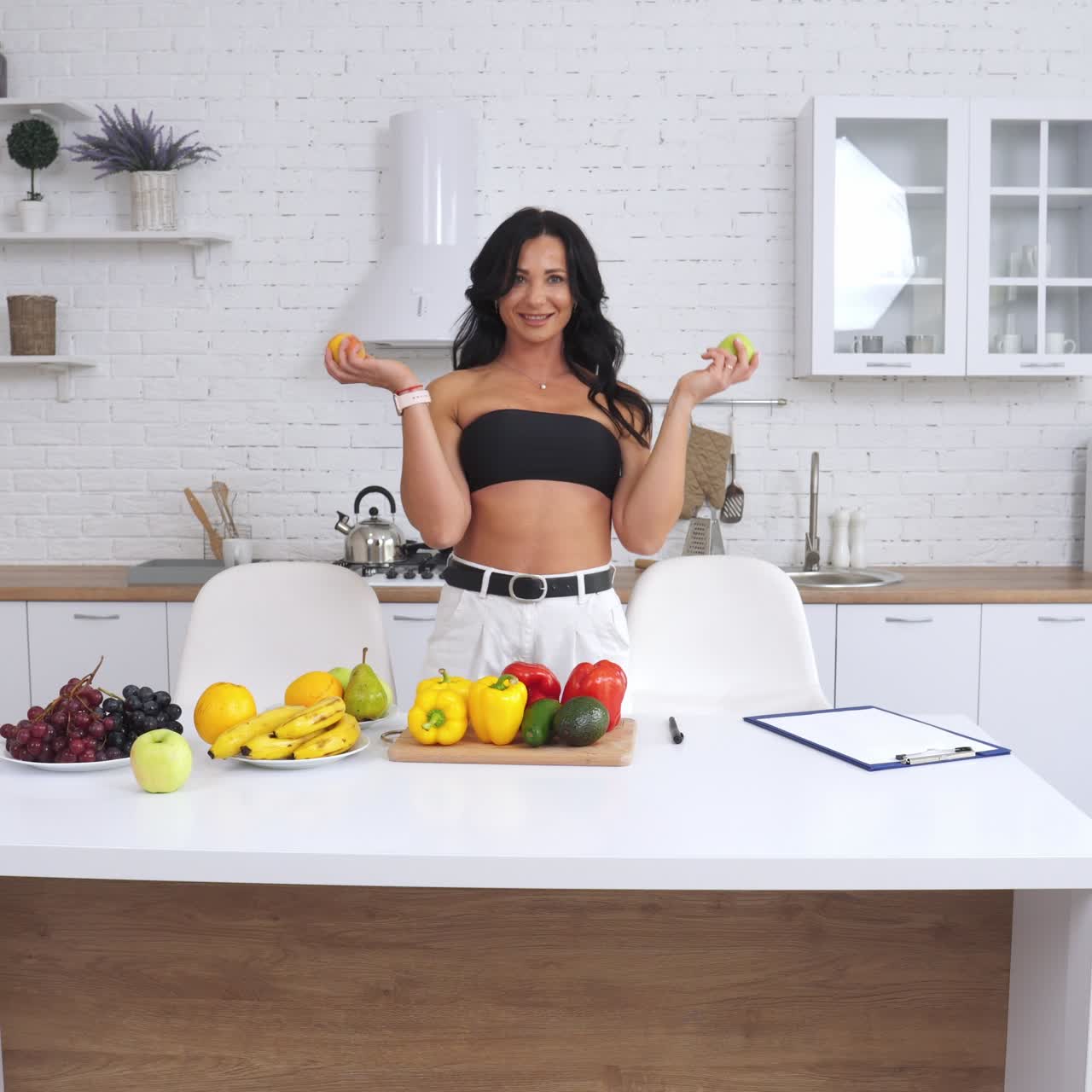 Attractive woman standing in light kitchen in front of the table with organic food. Lady proposes apple or a peach