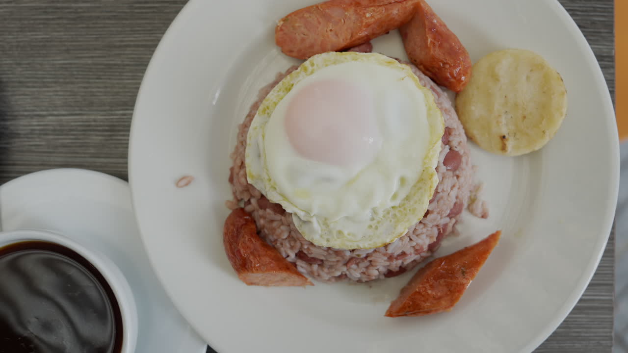 Enjoy a classic Colombian breakfast featuring calentado rice, fried egg, sausages, and arepa. Bright, inviting setup with freshly brewed coffee captures the essence of morning meals