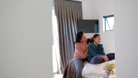 Happy-biracial-mother-talking-with-daughter-and-taking-care-of-her-hair-in-sunny-bedroom