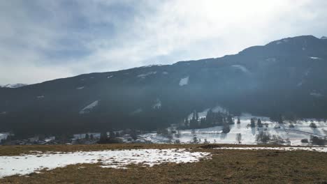 vista aérea de drones volando sobre una colina de campos de hierba marrón, revelando un camino con bosque y un pequeño pueblo a pie de una montaña alpina cubierta de nieve en un cálido y hermoso y soleado día de invierno con cielo azul