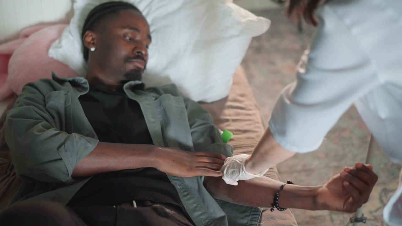 Home bedside emergency care paramedic attends patient, assessing arm for intravenous access, inserting cannula and securing dressing, nurse assisting with supplies, calm domestic atmosphere with soft