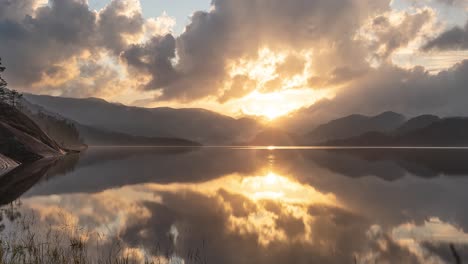 the setting sun shines through the heavy clouds above the calm lake in the nordic landscape