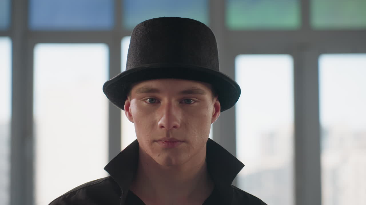 confident man in black shirt and hat tilting brim gazing at camera inside loft studio with large window and mirrored reflections showing neck tattoo under popped collar and subtle smile