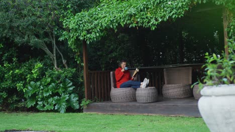 Ältere-Kaukasische-Frau-Entspannt-Sich-Im-Garten,-Sitzt-Mit-Erhobenen-Füßen,-Trinkt-Kaffee-Und-Liest-Ein-Buch