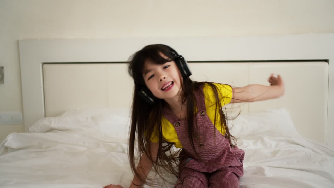 encantadora asiática escuchando música en el teléfono inteligente con auriculares en la cama y bailando divertido
