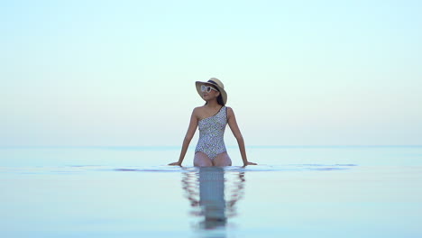 Asian-model-woman-sitting-at-the-edge-of-the-infinity-pool-leaning-on-hands-and-moving-her-legs-under-the-water-slow-motion-static,-copy-space-template