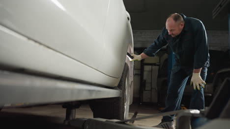 mechanic in blue uniform uses led lamp to inspect tire of a white car in garage, bending down with left hand on knee, showing focus and precision