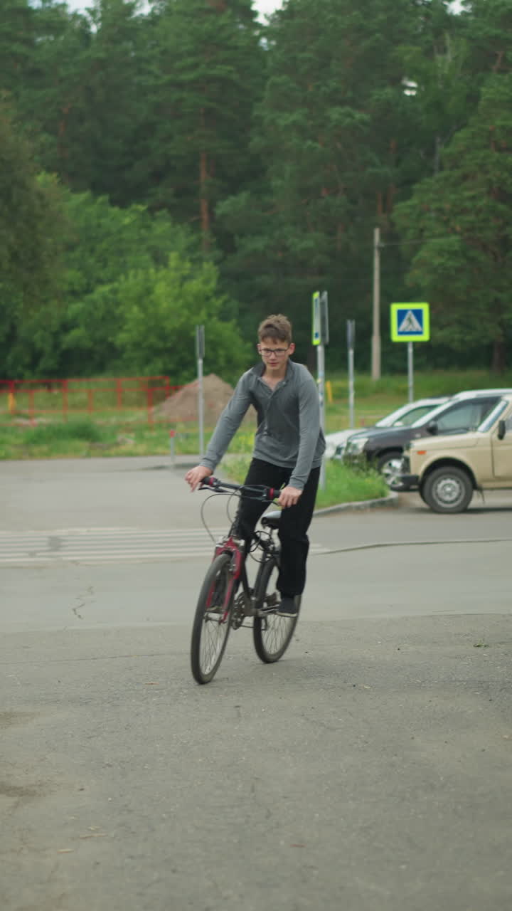 안경을 쓴 어린 소년은 초록색으로 둘러싸인 인터록 경로에서 자전거를 타고, 주차된 자동차, 보행자, 배경의 표지판, 조용한 교외 거리, 배경에 초록색이 있습니다.