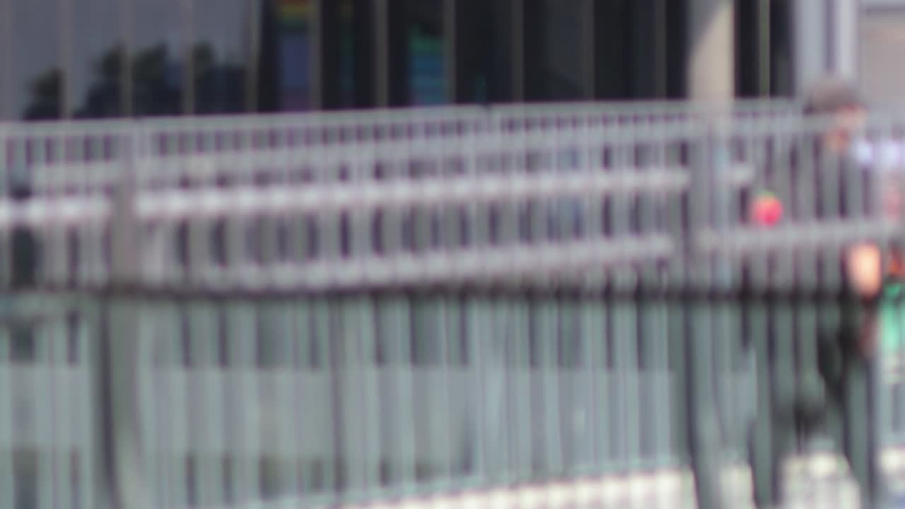 Blurred pedestrians move past a metal railing in an urban setting, with a glass building in the background.