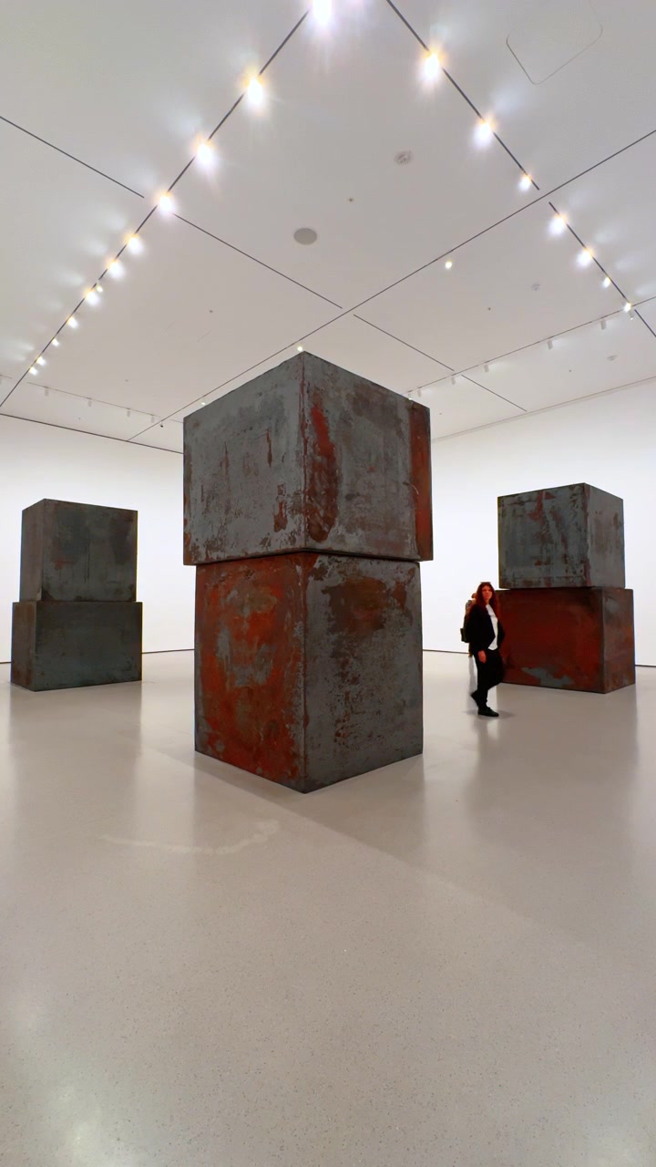 New York, USA, 12 agosto 2025: Scultura di cubo in acciaio arrugginito in una sala bianca. Un cubo metallico geometrico si erge sotto i riflettori mentre un visitatore scatta una foto
