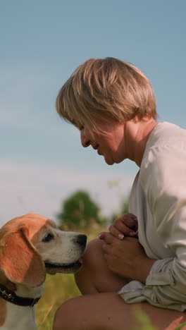 pet lover squatting outdoors with her dog in vast grassy field on sunny day, interacting playfully as the dog looks up with tongue out happily jumps up and kissed her, surrounded by lush nature