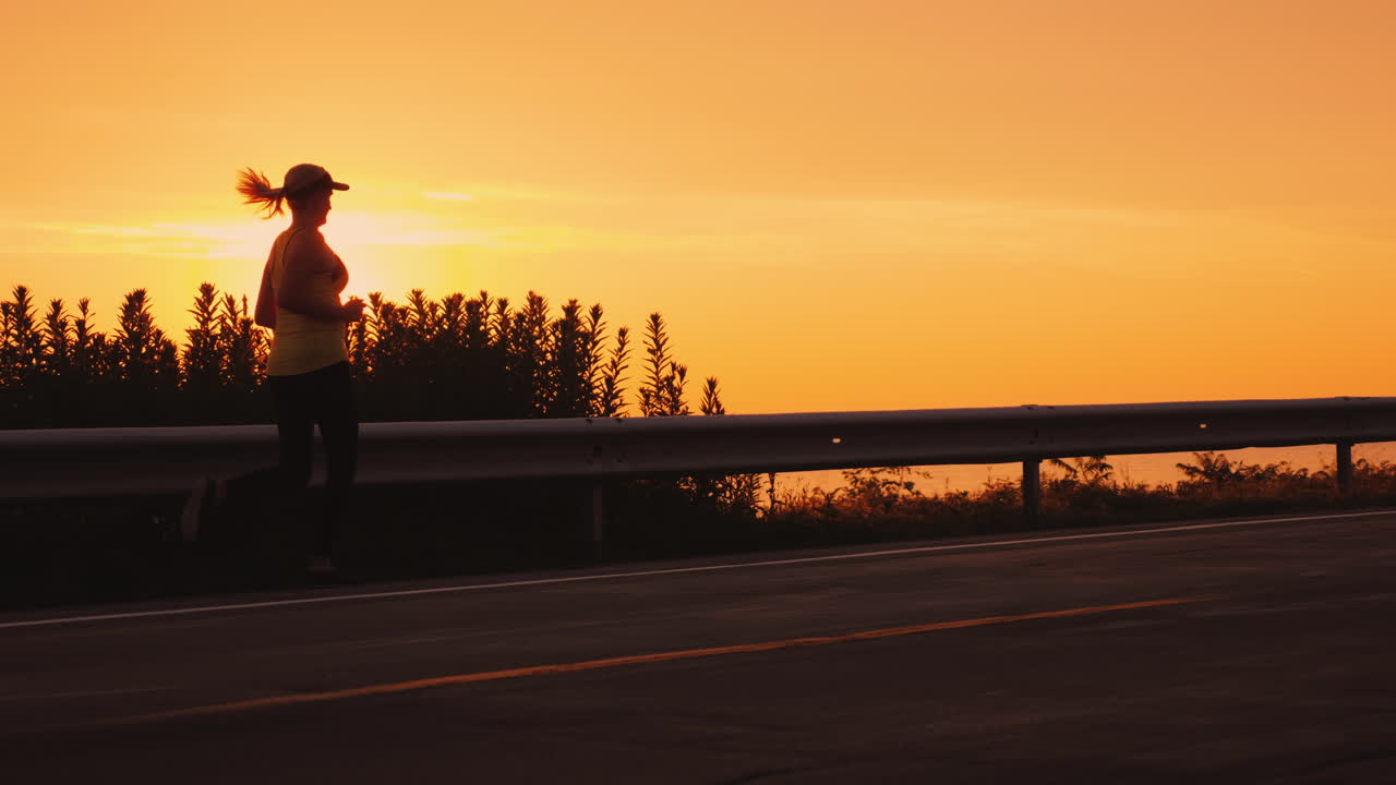 Slim Athletic Woman Running Along The Road Against The Orange Sky And The Setting Sun