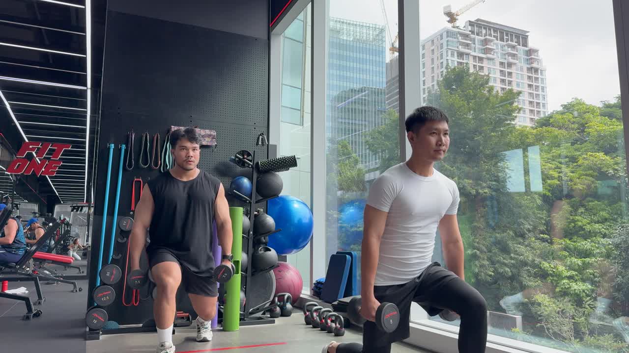 Two men exercise with dumbbells in a bright, modern gym. The environment is spacious with large windows and fitness equipment