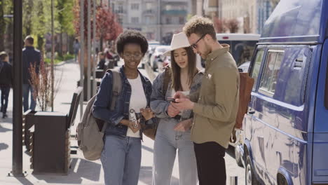 Three-Tourists-In-The-City,-Two-Young-Girls-And-A-Young-Male,-Looking-At-An-Address-On-The-Cell-Phone