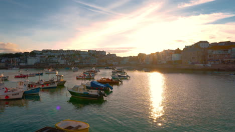 Famous-town-St-Ives-with-beach-and-harbor-promenade-in-Cornwall-in-Southern-England,-UK-with-fishing-boats-moving-in-the-clear-blue-water-by-sunset-and-reflections