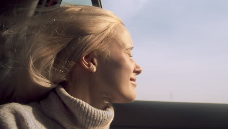 mujer sonríe por la ventanilla del coche abierta mientras el viento sopla pelo largo y rubio, primer plano