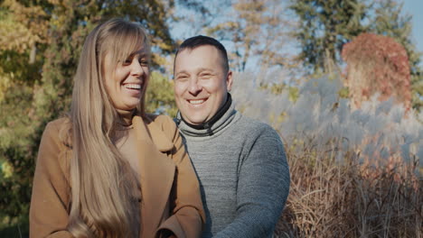 Retrato-De-Una-Pareja-Feliz-Sonriendo-Y-Mirando-A-La-Cámara.-De-Pie-Con-Ropa-Abrigada-En-El-Parque-De-Otoño