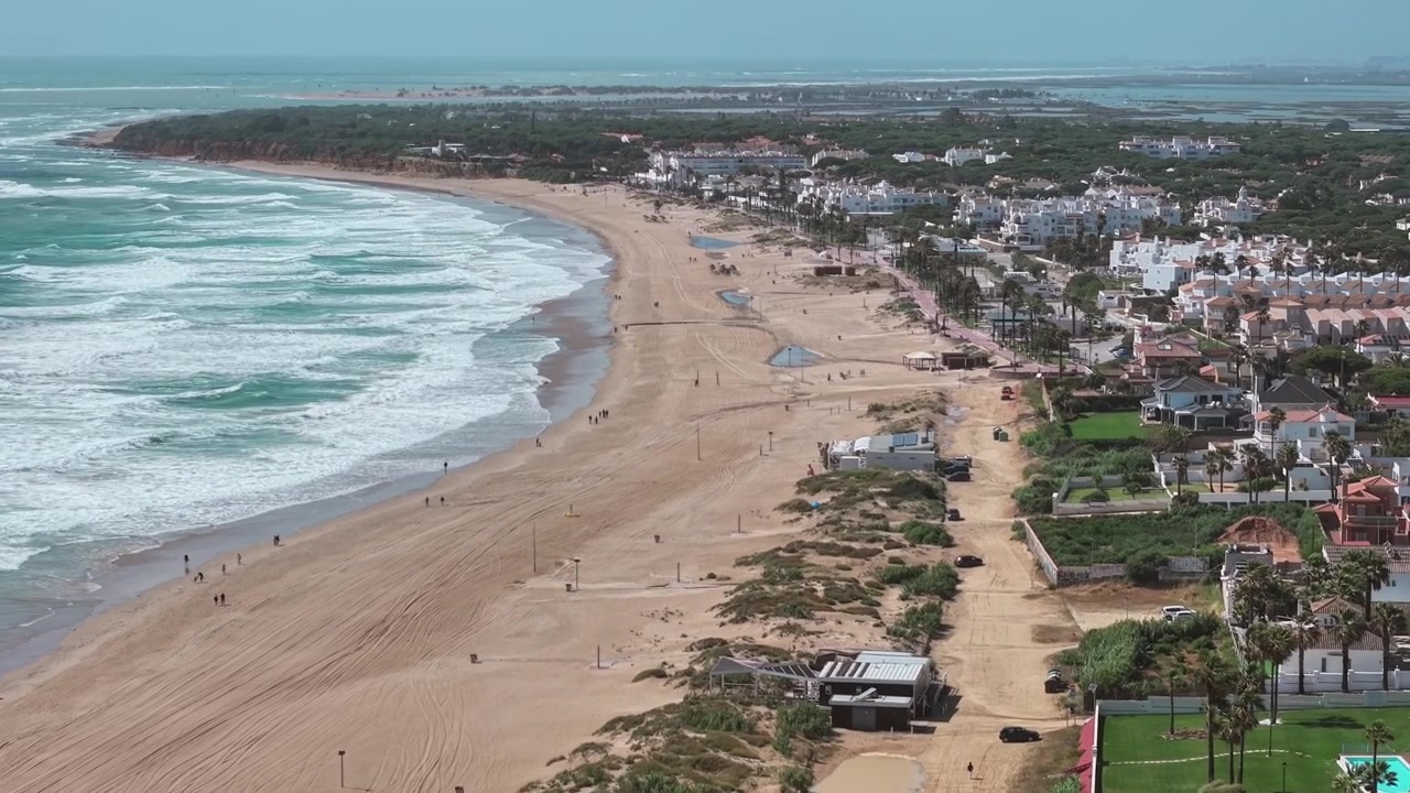Vuelo suave de dron hacia adelante sobre la costa de la playa de la Barrosa, que se extiende a lo largo de la zona residencial en Cádiz, Andalucía, España.