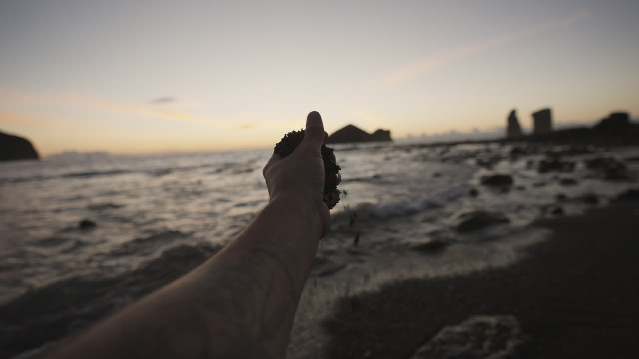 mano masculina recoge arena en la playa junto a la costa rocosa de las azores al atardecer, punto de vista