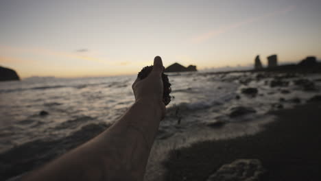 male hand picks up sand on beach by rocky azores coast at dusk, pov