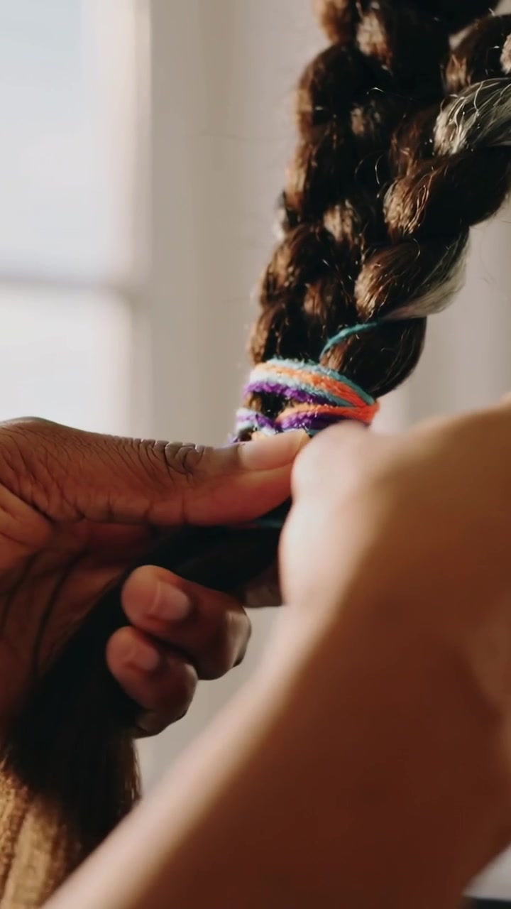 Vertical video: Approaching hands wrapping teal yarn around braid at home, securing colorful end