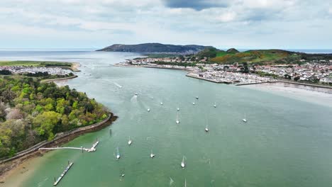 several sailboats anchor in the fast flowing conwy river with the coastal town of deganwy amongst the green hills of welsh nature on a cloudy day