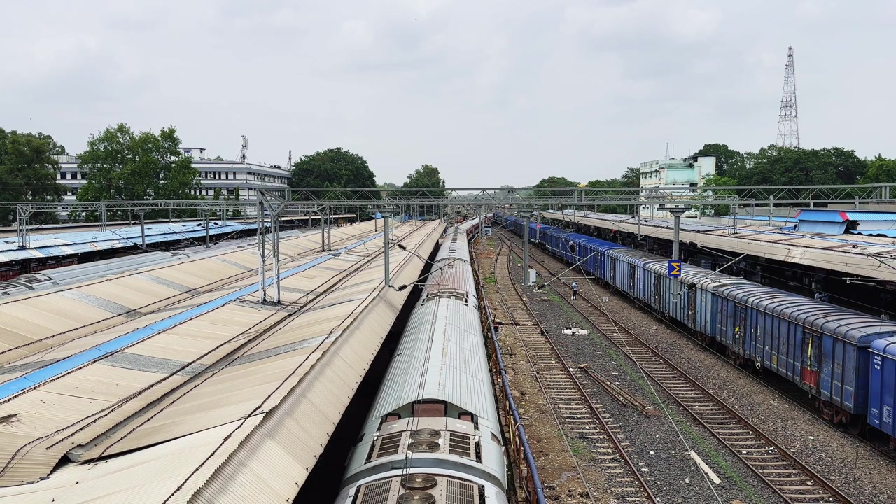 Inquadratura grandangolare di un treno passeggeri in partenza da una banchina ferroviaria con binari merci paralleli e linee elettriche aeree