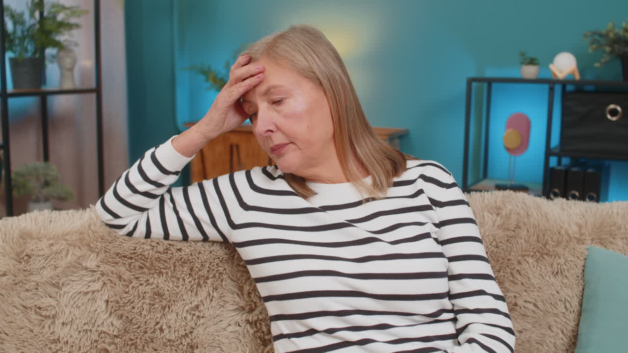 Mature woman placing hand on forehead on sofa showing frustration helplessness after bad day at home