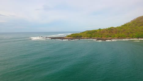 Pequeñas-Olas-Pasan-Bajo-Un-Dron-Que-Vuela-Hacia-Un-Punto-Rocoso-En-La-Costa-De-Costa-Rica