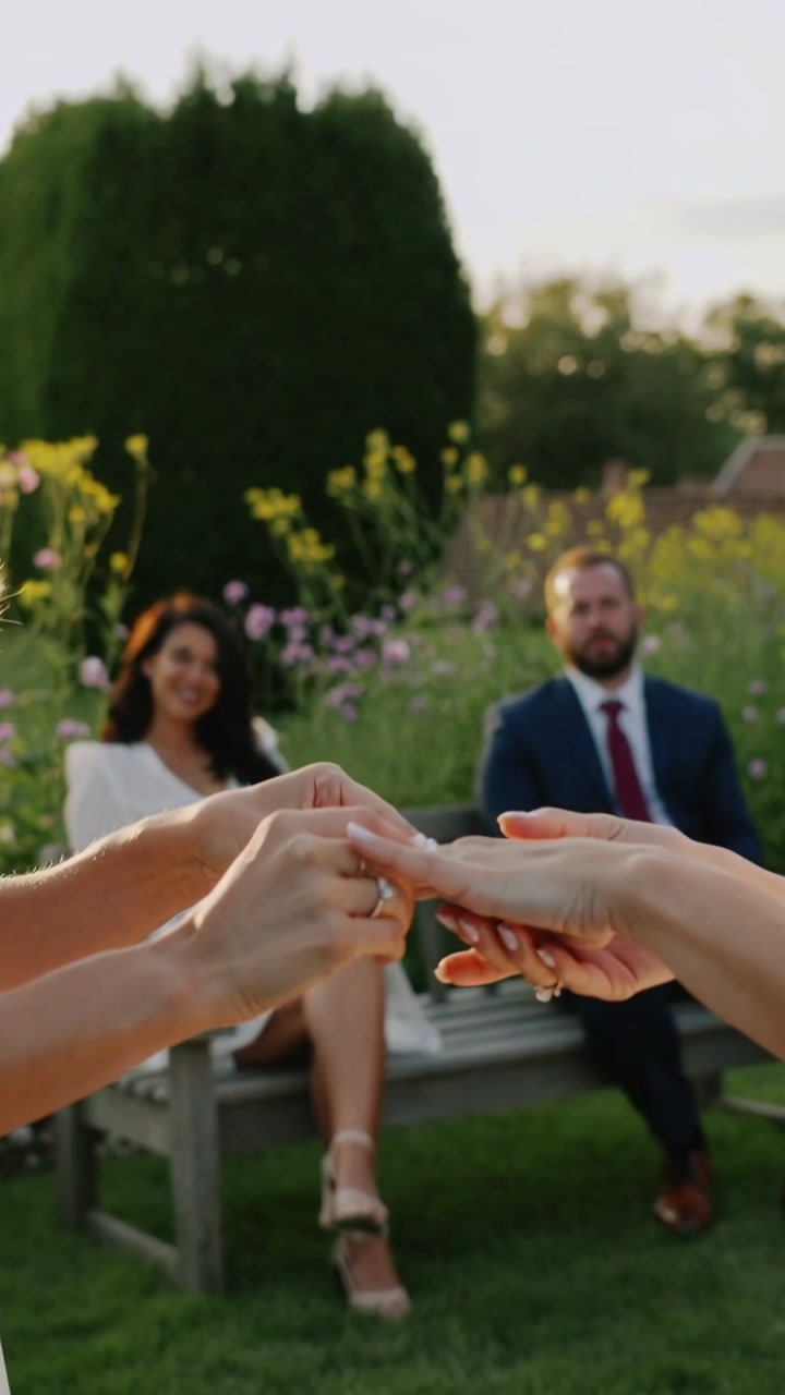 Vertical video: Partner placing wedding ring on partner's finger in garden during vows white dress