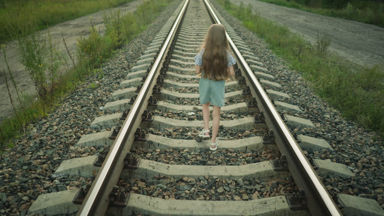 rear view of young girl with long hair walking alone on railroad track in rural setting, surrounded by grass and gravel, wearing striped shirt and denim dress in calm natural outdoor environment