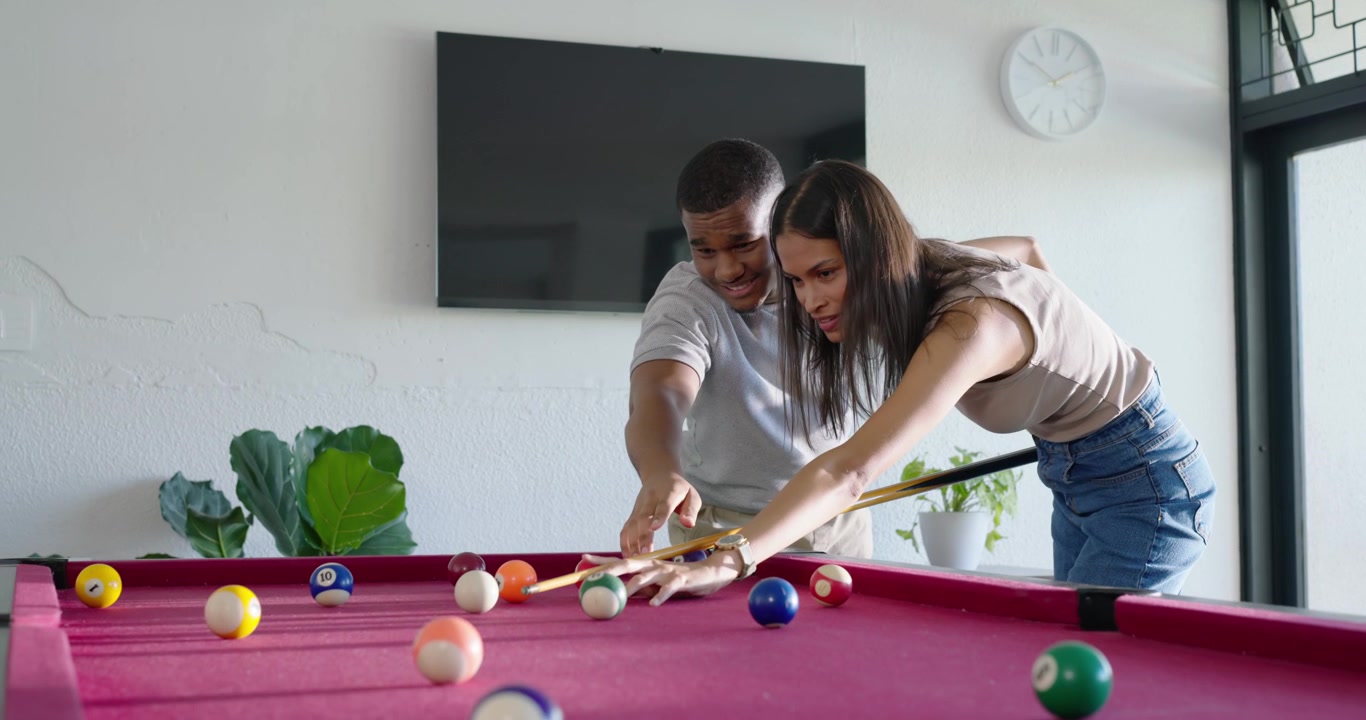 Pareja afroamericana jugando al billar en casa, el hombre guiando sus manos mientras ella hace el tiro, sonriendo.