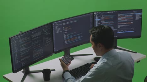 side view of happy asian man developer looking at code on mobile phone while write code with multiple computer screens in green screen studio
