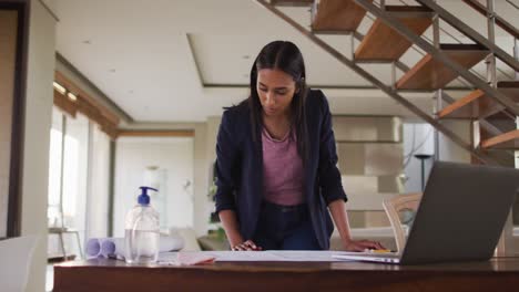 Mixed-race-woman-by-desk-at-home-using-laptop-looking-through-blueprints