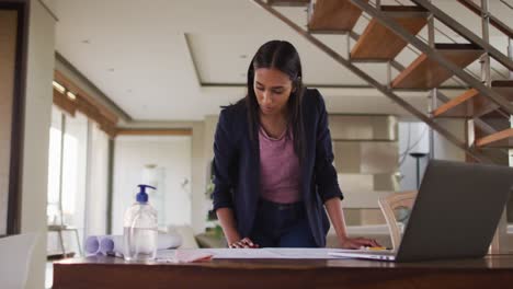 mujer de raza mixta en el escritorio en casa usando una computadora portátil mirando a través de planos