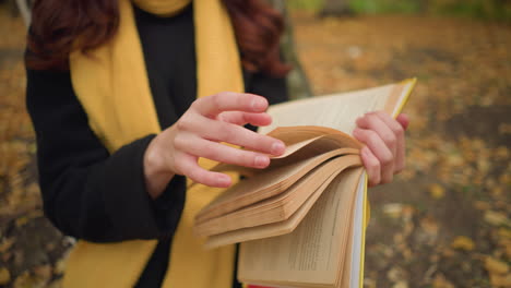 primer plano de la mano de la señora con una bufanda larga mientras sostiene el libro y hojea las páginas, hojas de otoño esparcidas por todas partes, creando una atmósfera de lectura cálida y pacífica en la naturaleza