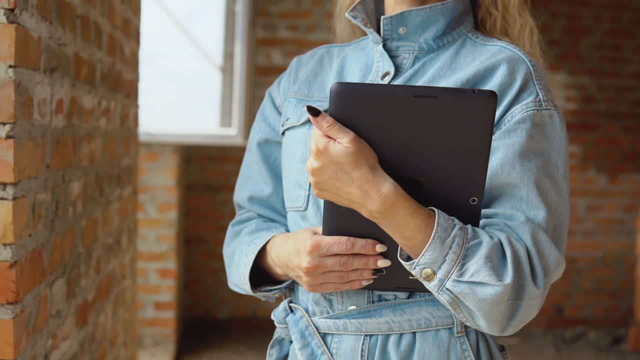 A female architect or bricklayer stands in a newly built house with untreated walls with a tablet in her hands. Modern technologies in the oldest professions
